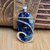 A handmade pendant featuring a polished blue and turquoise stone wrapped in silver-toned metal, displayed against a rustic brown leather and wood background.