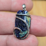 A close-up of a hand holding a silver-wrapped pendant featuring a polished blue and green stone with a bail at the top.