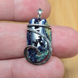 A close-up of a hand holding a small pendant made from a polished blue-green stone wrapped in silver wire, with a bail at the top.