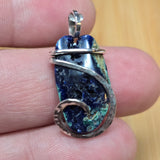 A close-up of a hand holding a small handmade pendant featuring a blue and green marbled stone wrapped in silver-toned metal.