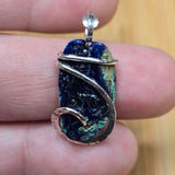 A handmade pendant featuring a textured dark blue lapis lazuli stone wrapped with silver and copper wire, held on a person's fingers.