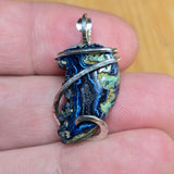 A handmade silver wire-wrapped pendant featuring a blue and green agate stone, held in a person's hand against a wooden background.