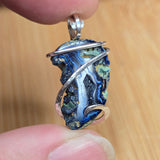 A close-up of a handmade pendant featuring a blue and green agate-like stone wrapped in silver wire, held between two fingers against a light wood background.