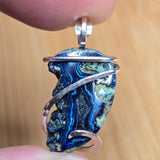A close-up of a hand holding a handmade pendant featuring a polished blue agate stone wrapped in silver-colored metal.