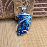 A close-up of a handmade pendant featuring a blue and green agate stone wrapped in silver-colored metal, resting against a brown leather strap on a wooden surface.