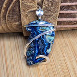 A close-up of a handmade pendant featuring a polished blue and green agate slice wrapped in silver-toned metal, resting on a brown leather strap over a light wood surface.