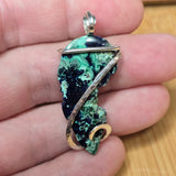 A handmade pendant featuring a green and black textured stone wrapped in silver and gold wire, held in a person's hand.