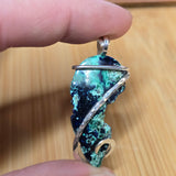 A close-up of a hand holding a handmade pendant featuring a marbled dark blue and green stone wrapped in silver-toned metal.