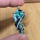 A close-up of a hand holding a raw, polished gemstone pendant with a silver and gold wire wrap, set against a light wood grain background.