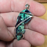 A handmade pendant featuring a green and black mottled stone wrapped in silver-toned metal, held in a person's hand.