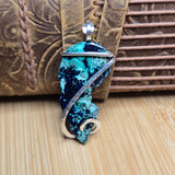 A handmade pendant featuring a turquoise and dark blue stone with silver swirls, resting on a brown leather strap over a bamboo surface.