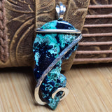 A handmade pendant featuring a turquoise and dark blue marbled stone cabochon wrapped in a hammered silver wire bail, displayed against a brown leather strap and wooden background.