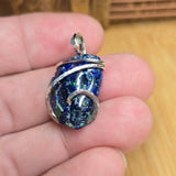 A handmade silver-wrapped pendant featuring a textured, iridescent blue and green stone, held in a person's hand against a blurred wooden background.