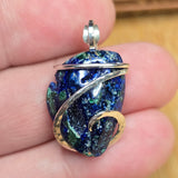 A close-up of a hand holding a small pendant made from a polished blue and green stone with silver and gold wire wrapping and a silver bail.
