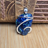 A blue and green metallic stone pendant wrapped in silver-toned metal, displayed on a wooden surface in front of a brown leather strap.