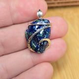 A handmade glass pendant with a deep blue and green marbled pattern, wrapped in silver wire, held in a person's hand.