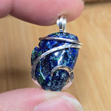 A close-up of a hand holding a small pendant made from a blue and green agate stone wrapped with silver wire, against a light wood background.