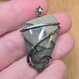 Gray blue greenstone pendant wrapped in a swirl of sterling silver held on fingertips against blurred background. 