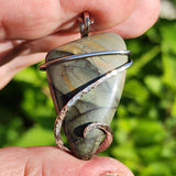 Gray blue greenstone pendant wrapped in a swirl of sterling silver held on fingertips against blurred green background. 