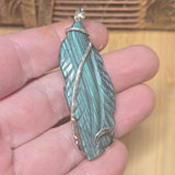 Teal feather-shaped pendant held in a hand with a wooden background