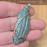 Teal feather-shaped pendant with silver wire wrapping held in a hand against a wooden background