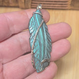Turquoise labradorite feather-shaped pendant held in a hand on a wooden surface