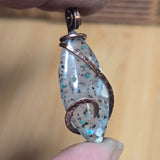 Hand holding a decorative pendant with speckled design and copper wire wrapping against a blurred background.
