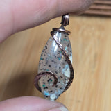 Close-up of a hand holding a decorative pendant with speckled material and copper wire wrapping.