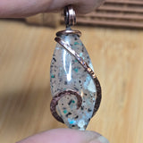 Marquis-shaped pendant with decorative copper wire wrapping held between two fingers on a wooden surface.