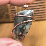 Pendant with a stone and copper wire wrapping held between fingers against a wooden background