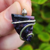 Hand holding a purple stone pendant with silver wire wrapping against a green leafy background