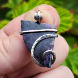 Hand holding a purple stone pendant with silver wire wrapping against a green leafy background