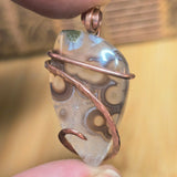 Pendant with a tube agate stone and copper wire wrapping held between fingers against a blurred background