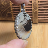 Close-up of a silver pendant with a natural stone design held by a hand against a wooden background.