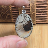 Oval-shaped pendant with natural stone and silver frame held by a hand on a wooden surface