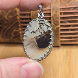 Oval-shaped stone pendant with silver frame held between fingers against a wooden background