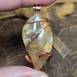Oregon Plume agate in copper wire wrap held between two fingers in front of blurred brown pattern background