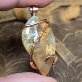 Plume Agate pendant with copper wire wrapping held between fingers against a textured brown background