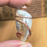 Hand holding a pendant with a clear stone and copper wire wrapping against a blurred background