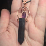 Dark fluorite stone pendant with purple gemstone on a copper chain held in a hand.