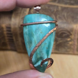 Green stone pendant with copper wire wrapping held by a hand against a wooden background