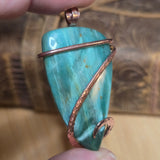 Green stone pendant with copper wire wrapping held in hand against a blurred background