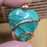 Heart-shaped green jasper pendant with copper accents held between fingers against a blurred background