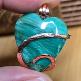 Turquoise green heart-shaped pendant with copper accents held between fingers on a wooden surface