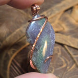 Hand holding a purple stone pendant with copper wire wrapping against a blurred natural background