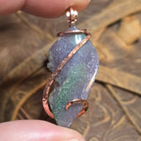 Hand holding a pendant with a purple stone and copper wire wrapping against a blurred natural background