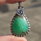Green pendant with silver spiral design held between fingers against a blurred background