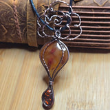 A handmade wire-wrapped pendant necklace featuring a teardrop-shaped amber stone with red inclusions, hanging from a dark cord with an ornate metal top, set against a wooden background.