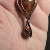 A close-up of a teardrop-shaped pendant with amber-colored stones and a dark metal frame, held in a person's hand.