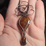 A close-up of a hand holding a handmade pendant necklace. The pendant features a teardrop-shaped amber stone with a smaller amber teardrop at the bottom, set in silver-toned metal with a decorative wire loop at the top.